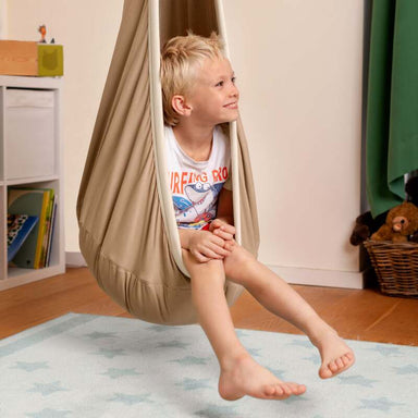 LA SIESTA Joki Teddy Kids Hanging Nest – kid seated beside a playroom bookshelf